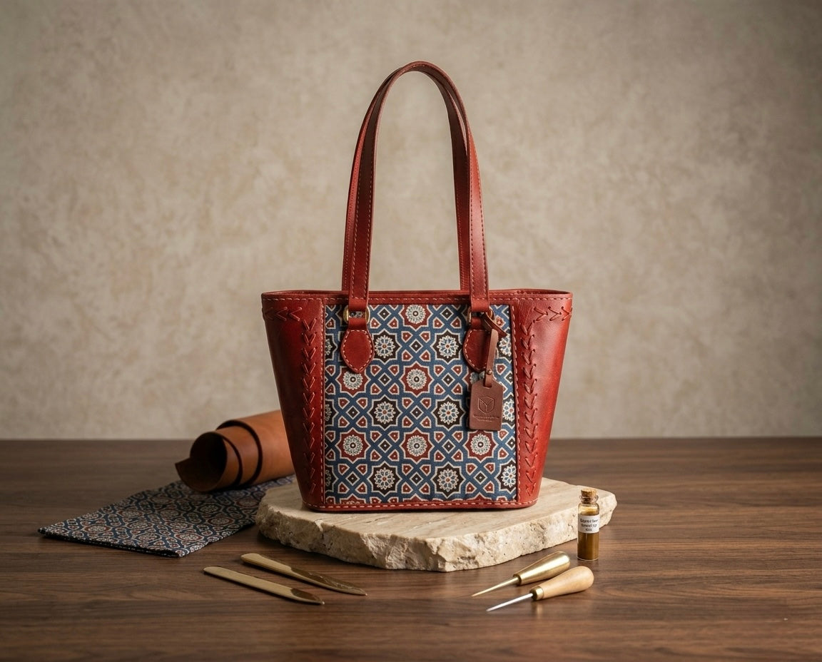 A leather tote bag with a geometric patterned front panel, displayed on a stone slab on a wooden table with leatherworking tools and a rolled leather piece in the background.