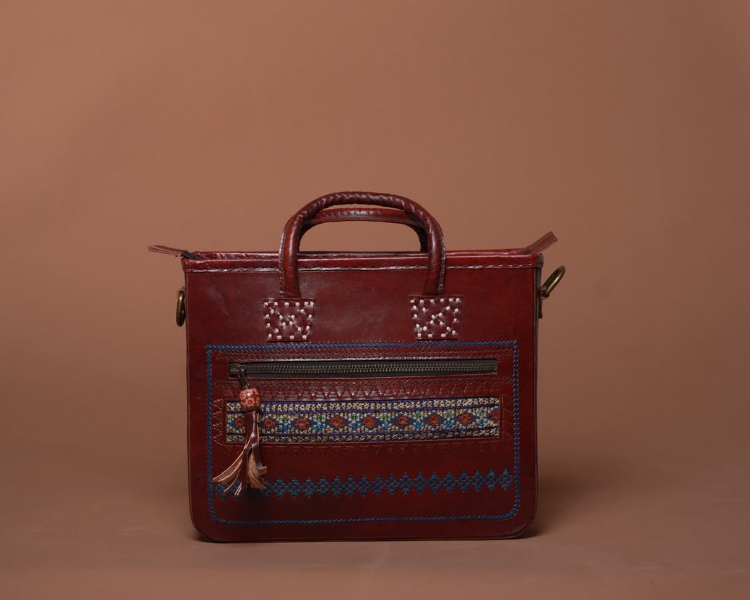 A dark red leather handbag with a top handle, decorative geometric embroidery on the front pocket, and a small hanging tassel charm, set against a solid warm brown background.