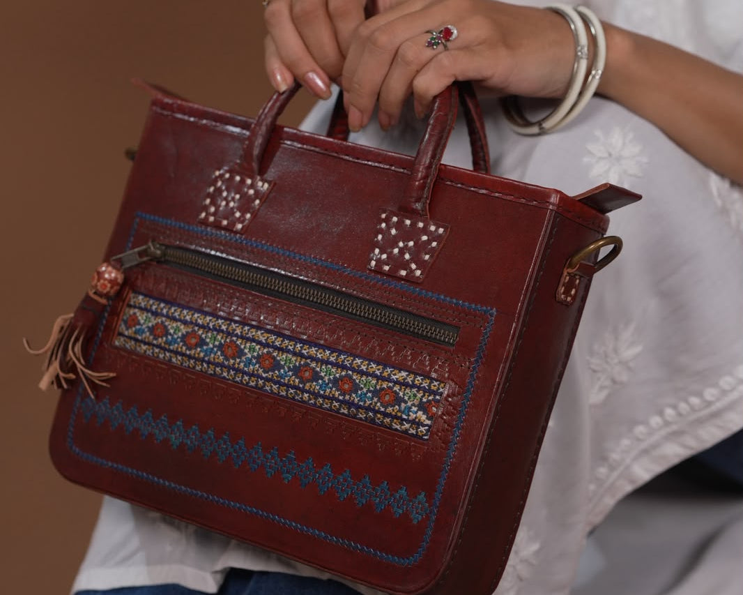 A person holding a small reddish-brown leather handbag with a geometric embroidered pattern across the front, paired with a white embroidered top and denim skirt against a warm brown background.