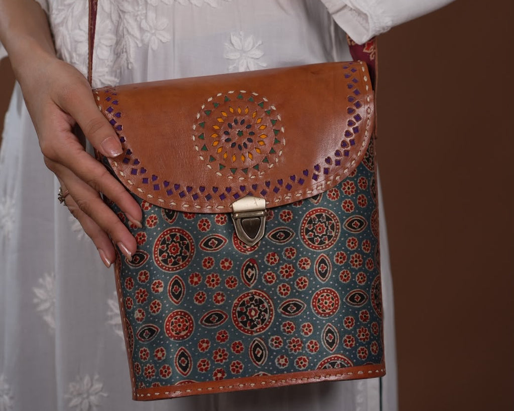 A person in a white embroidered dress holds a brown leather crossbody bag with a colorful patterned body and a circular mandala design on the flap.